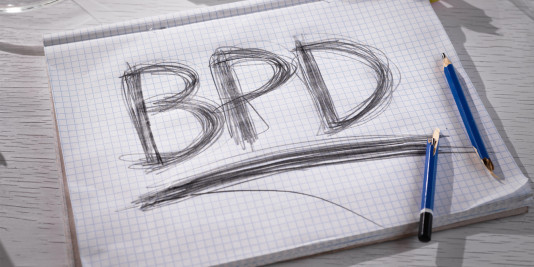 The abbreviation for borderline personality disorder (BPD) is expressively written in pencil with strong pressure on paper. There is crumpled paper and a broken glass on the table. Strong emotional experience.