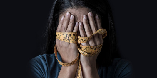 Close up hands wrapped in tailor’s measuring tape covering face of young woman with an eating disorder against a black background.